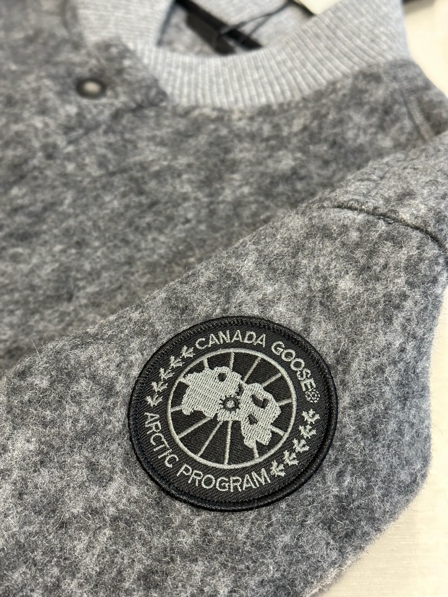 P770 Canada Goose Fleece Badge Button Jacket The main fabric of this jacket is light gray wool blend fabric to improve the fabric's wear resistance, wrinkle resistance and warmth. Wool provides good warmth and a natural feel, while blended fibers enhance the fabric's practicality and durability.  The cutting process needs to ensure that the texture and color of the fabric are evenly distributed, especially with this light gray tone, to avoid chromatic aberrations. The stretchability of wool-blend fabrics needs to be taken into consideration when cutting pieces to ensure dimensional stability of the finished garment.  Use a sewing needle and thread suitable for wool-blend fabrics to avoid damage to the fabric during sewing. The sewing stitches are even and firm to prevent thread stripping.  The temperature needs to be controlled during ironing to avoid high temperature damage to the fabric while keeping the fabric smooth and crisp. The front and back body parts, sleeves and other main parts are stitched with flat seams to ensure a smooth silhouette. Align the fabric texture at the splices to keep the look neat.  The white circular armband on the left arm is an important part of the brand identity and is finely sewn to ensure it is strong and flat.  size：s m l xl